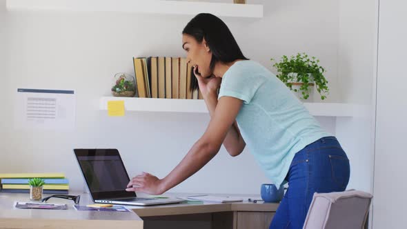 Mixed race gender fluid person talking on smartphone working from home using a laptop