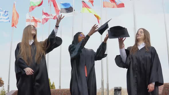 Three Young Women of Different Nationalities Joyfully Throw Hats Up Against Many Flags of Different