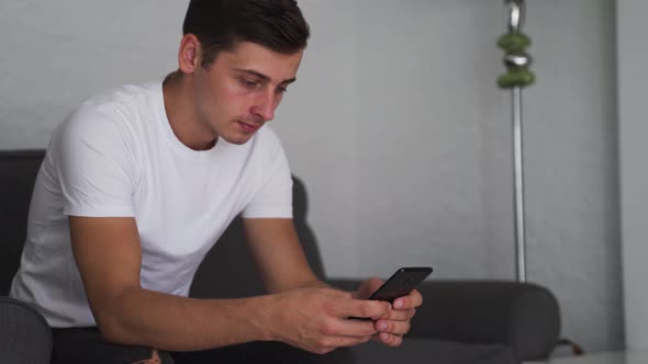 Happy young man uses a smartphone while sitting on a sofa. Browse online internet, shopping and use