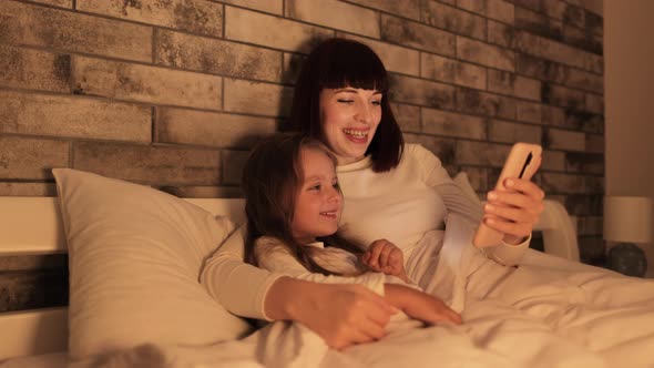 Mother and Her Little Happy Daughter Lying in Bed with Smartphone Covered with Blanket