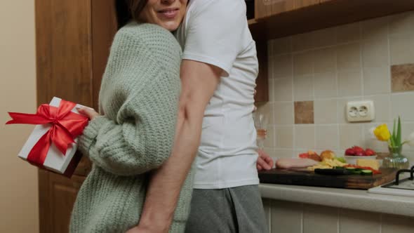 Young Woman Giving Gift To Her Husband Who Prepares Food in the Kitchen