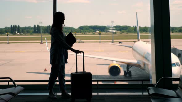 Dark Silhouette of Businesswoman with Rolling Bag and Laptop in Her Hands, Standing Against