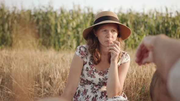 A Serene Mother in a Summer Dress and a Straw Hat Eats and Watches Her Children at the Family