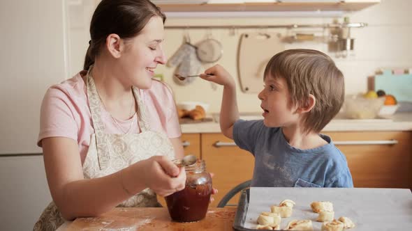 Smiling Little Boy Feeding His Mother with Jam From Spoon While Making Biscuits at Home