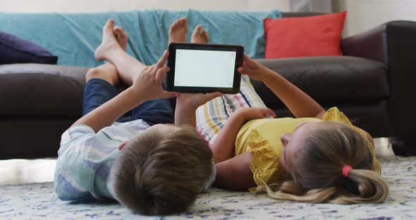 Happy caucasian brother and sister lying on floor in living room using tablet, copy space on screen