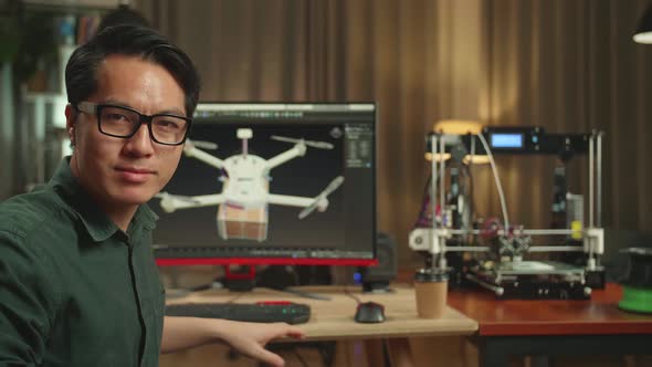 Asian Engineer Work On Personal Computer And 3D Printer. He Turns And Warmly Smiles Into The Camera