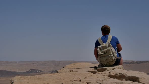 Young Man with Backpack Sitting on Cliff's Edge and Looking at the Desert