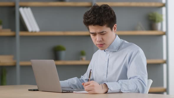 Young Asian Man Lefthander Watching Online Lesson on Laptop and Taking Notes Studying Distantly on