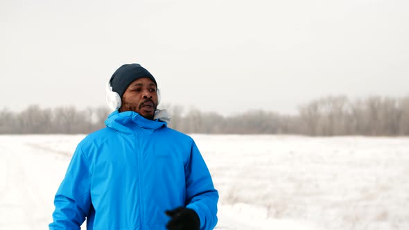 Black Man in Warm Clothes and Headphone Runs Along Road in Winter Outdoors Stops and Catches Breath