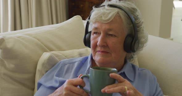 Caucasian senior woman wearing headphones listening to music while holding coffee cup at home