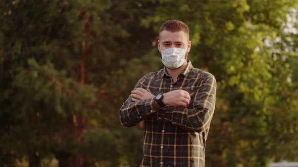 Confident Hipster Man in Medical Mask Stands Against Background of Green Trees