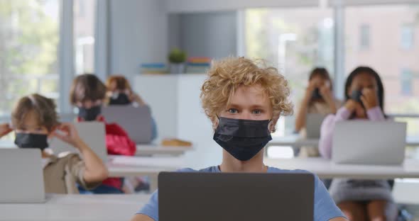 Portrait of Diverse Children Put on Face Mask Sitting at Desk in Class Looking at Camera