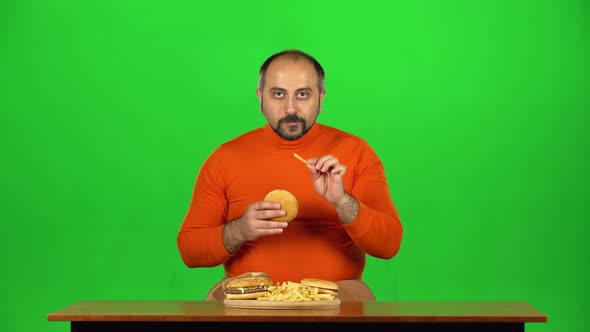 Man at a Table with a Plate of Fast Food Enjoys Eating a Burger and French Fries, Green Screen