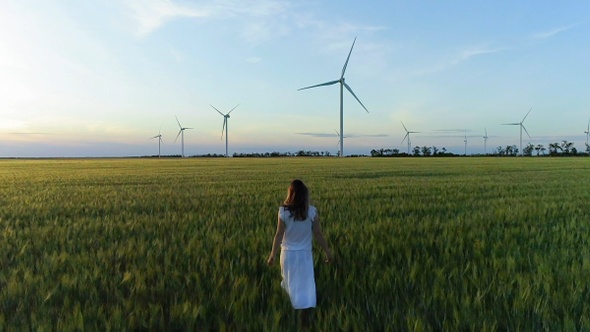 Beautiful girl walking on a green wheat field with windmills for electric power production