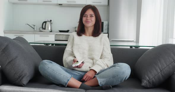 Cute Female Watches Boring Movie on Tv and Switches Channels on Remote Control Sits Crossed Legs on