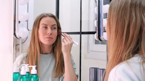 Young Woman Applying Powder with Make Up Brush to Face Looking to Mirror