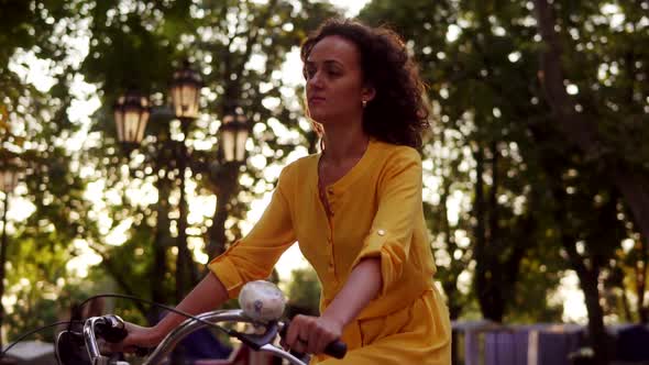 Lens Flare Young Woman in Long Yellow Dress is Riding a City Bicycle with a Basket and Flowers in