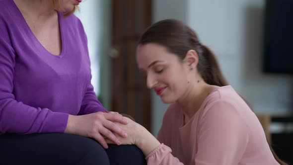 Frustrated Caucasian Young Woman Crying Kissing Hands of Unrecognizable Mother Putting Head on Knees