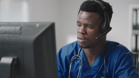 A Black Man Sits at a Computer in a Doctor's Uniform and Writes a Patient's Card While Taking Calls