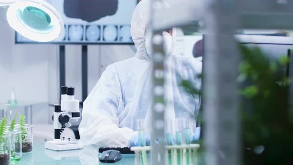 Biologist Dressed in Protective Coverall and Wearing a Mask Is Lookign at a Microscope