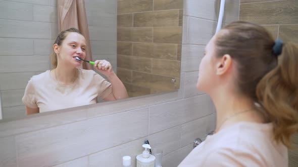 Woman in Beige Tshirt Actively Brushes Her Teeth with Green Toothbrush in the Bathroom at Home