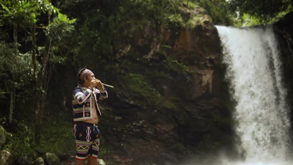 young man playing bamboo flute behind is a waterfall while standing