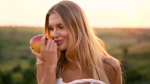 Beautiful sexy blonde girl in white dress posing in a field at sunset with a basket of fruit