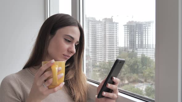 Young Woman Smiling Drinks Coffee and Using Smartphone Surfing News or Chatting Near Window