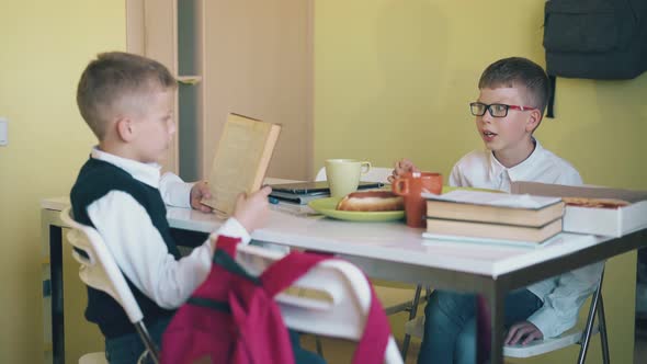 Schoolboy Reads Book To Friend Eating at Table in Canteen