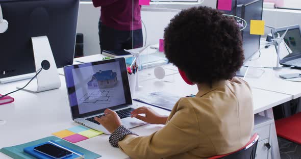 Mixed race businesswoman wearing face mask sitting using a laptop in modern office