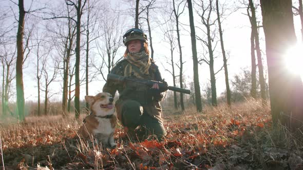 Young Pretty Redhead Woman in Military Uniform Armed with Rifle Playing with Dog