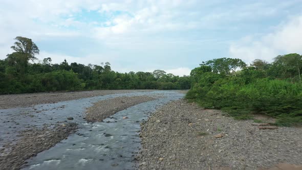 Drone shot flying over a tropical stream covered in pebbles and a lush green colored forest