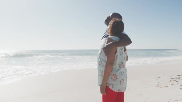 Smiling senior african american couple embracing and looking at sea on sunny beach