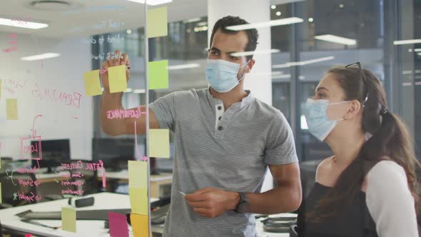 Diverse male and female work colleagues wearing face masks brainstorming using glass wall