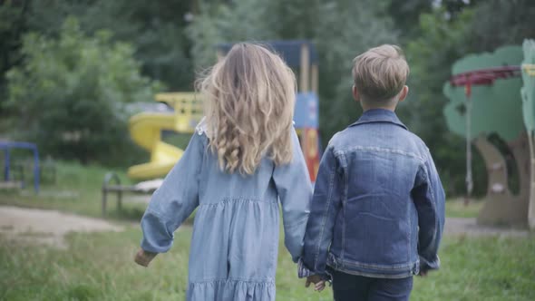 Back View of Little Caucasian Boy and Girl Holding Hands and Walking To Playground