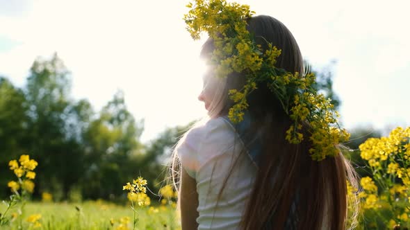 A Little Cute Girl with a Wreath of Yellow Flowers on Her Head Sits on a Sunny Flower Meadow