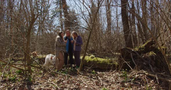 three girls stand forest holding a phone and a thermos in their hands, a white dog takes a stick