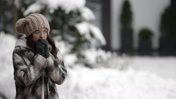 A Happy Young Darkhaired Woman in Round Glasses and a Plaid Coat Walks on a Snowy Winter Day Rubbing