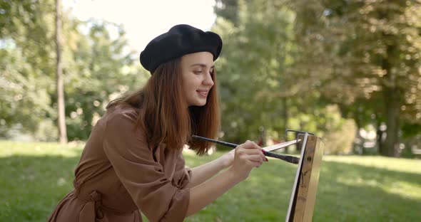 Girl Artist in a Beret Paints with a Brush on an Easel in the Park