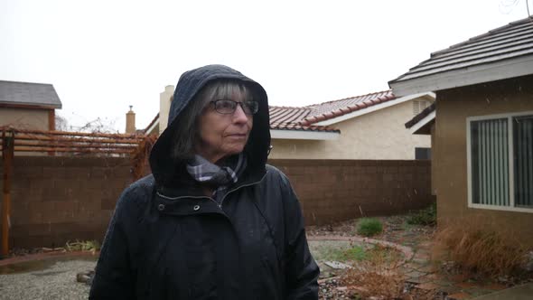 An elderly woman with glasses and a raincoat standing in a winter weather rain storm as raindrops fa