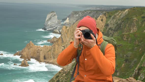 Man in Hat Takes Picture with Camera on Cliff Against Sea