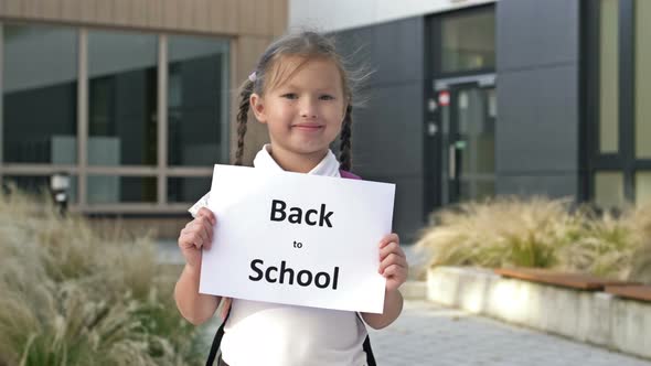 Cute Smiling Little Schoolgirl in Uniform Stands with a Poster Back To School Near the School