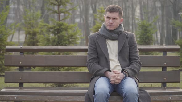 Nervous Caucasian Young Man Sitting on Bench in City Park and Checking Time on Watch. Portrait of