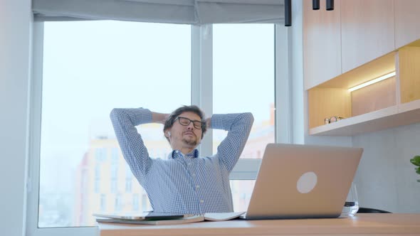 Young man is stretching at the computer, raising his hands up, resting from the work done.