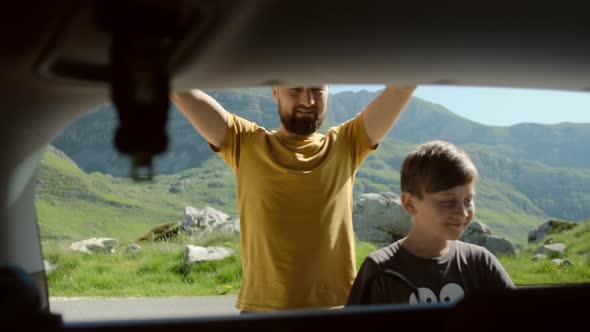 Happy father and son open car trunk and take backpack gets things out against mountain landscape