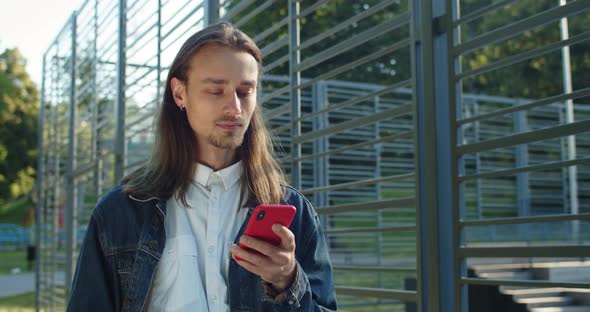 Crop View of Long Haired Guy Using Smartphone While Walking Near Sports Ground. Millennial Man with