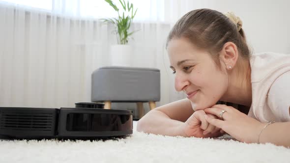 Portrait of Smiling Young Woman Looking at Working Robot Vacuum Cleaner in House