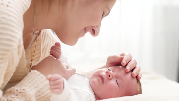 Portrait of Adorable Smiling Newborn Baby and Happy Young Mother Lying on Bed Against Big Window in