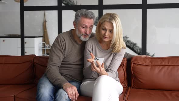 Hilarious Mature Couple Sitting on the Sofa and Holding Smartphone