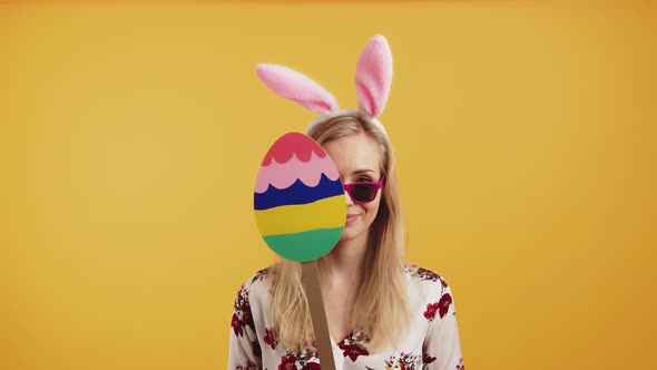 Humorous Caucasian Girl in Her 20s with Bunny Ears Cardboard Easter Egg and Sunglasses Posing at